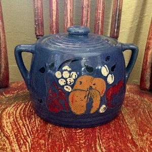 May include: Blue ceramic cookie jar with a lid and two handles. The jar is decorated with a hand-painted design of fruit and grapes in shades of orange, red, white, and black.
