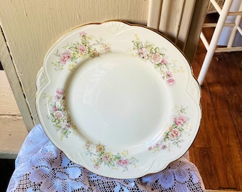 May include: Cream-colored ceramic plate with a scalloped edge and a gold rim. The plate is decorated with clusters of pink, yellow, and green floral designs. The plate is sitting on a white lace tablecloth.