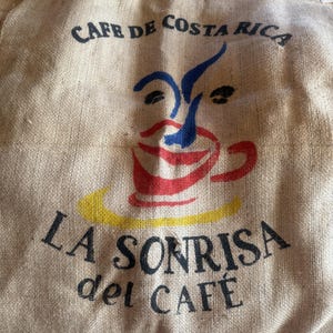May include: A tan burlap sack featuring the black text CAFE DE COSTA RICA and LA SONRISA del CAFE. A graphic print depicts a stylized face with a red coffee cup as the mouth, blue abstract lines for eyes and nose, and a yellow saucer base.