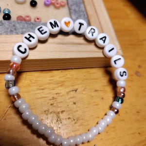 May include: A white beaded bracelet with the word "CHEMTRAILS" spelled out in black letters. A small orange heart bead is between the words "CHEM" and "TRAILS".