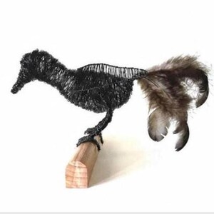 May include: A handcrafted wire bird sculpture with a black body and a tail of brown and gray feathers. The bird is perched on a small, light-colored wooden block, creating a unique decorative piece.