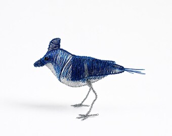 May include: A bird sculpture crafted from thin blue and silver wire. The bird features a prominent crest on its head, a dark eye, and a pointed beak. The body is primarily blue wire with a silver wire underbelly and thin silver wire legs.
