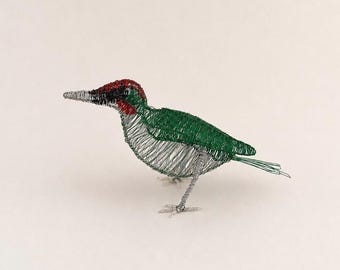 May include: A bird sculpture crafted from thin silver, green, and red wire. The bird features a silver wire beak, a red crown, a green patch near the eye, a green back and tail, and a silver wire chest and legs, standing on two wire feet.