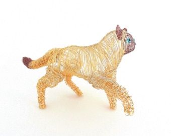 May include: A wire sculpture of a cat in a walking pose. The cat is made of gold-colored wire, with a brown tail and ears. The cat has blue eyes and is set against a white background.