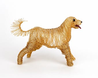 May include: A decorative sculpture of a dog crafted from thin gold-toned wire. The figure features a curled tail, four legs, and a head with small black bead eyes and a dark red beaded tongue protruding from the mouth, all set against a white background.