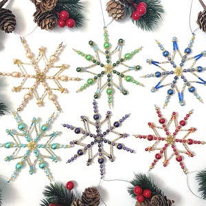 May include: Six beaded snowflake ornaments in various colors, including gold, green, blue, purple, turquoise, and red. Each snowflake has a central gold bead and radiating arms. The ornaments are arranged on a white surface with pinecones and red berries.