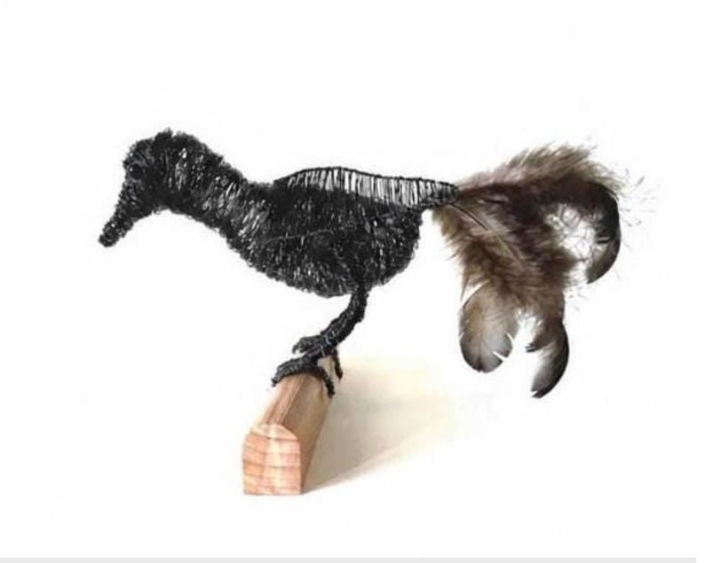 May include: A handcrafted wire bird sculpture with a black body and a tail of brown and gray feathers. The bird is perched on a small, light-colored wooden block, creating a unique decorative piece.