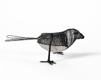 May include: A bird sculpture crafted from thin black and silver wire. The bird features a dark head and back with a contrasting silver-toned wire underbelly, thin wire legs, and a long, slender tail made of looped black wire strands.