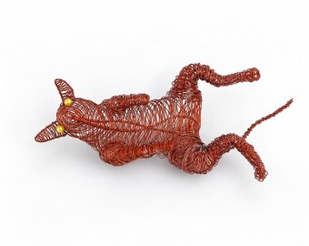 May include: A sculpture of a cat crafted from coiled copper-colored wire. The feline figure is depicted in a reclining position with its limbs extended. It features two small, round, yellow beads serving as eyes, integrated into the wire structure.