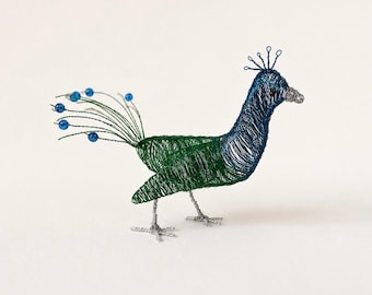 May include: A decorative peacock sculpture crafted from thin blue and green wire. The bird features a blue head with a wire crest, a green body, and a tail made of wire strands tipped with small blue beads. It stands on two silver wire legs.