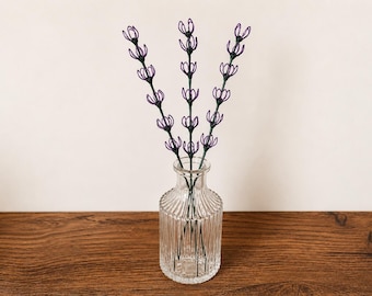 May include: Three decorative stems featuring purple wire-formed flower shapes arranged on dark green stalks. The stems are placed inside a clear, vertically ribbed glass vase with a narrow neck and a wider, rounded base.