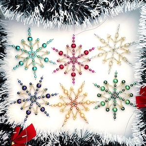 May include: Six beaded snowflake decorations in shades of teal, pink, gold, purple, yellow, and green. Each snowflake is crafted with beads and wire, ideal for festive displays. The background features tinsel and red bows, adding to the holiday theme.