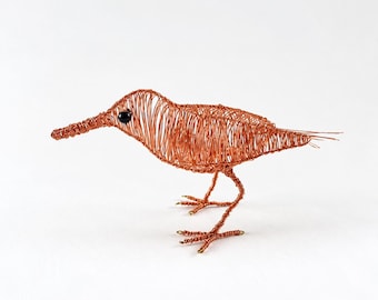 May include: A sculpture of a bird crafted from thin copper wire. The figure features a long, slender beak, a rounded body constructed from coiled wire strands, and two thin legs with detailed feet. A single small black bead serves as the eye.