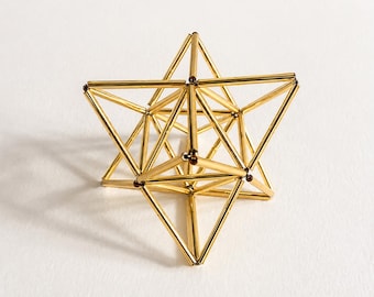 May include: A geometric sculpture constructed from thin, shiny gold-toned metal rods connected at dark brown spherical joints. The structure forms a complex three-dimensional star shape with multiple triangular faces and pointed vertices.