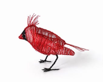 May include: A sculpture of a cardinal bird crafted from thin red wire, featuring a prominent crest on its head, a black wire beak area, and two black wire legs. The body is constructed with a dense, layered wire mesh technique.
