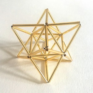 Puede incluir: Una escultura geométrica de estrella dorada. La escultura está compuesta por elementos triangulares y lineales interconectados, creando una forma de estrella tridimensional. La estructura metálica se encuentra sobre un fondo blanco.