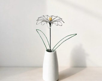 May include: A silver wire flower with a small gold-colored center bead, featuring a dark green wire stem and two green wire leaves, displayed in a smooth, tapered white ceramic vase. The flower has multiple looped petals made of thin silver wire.