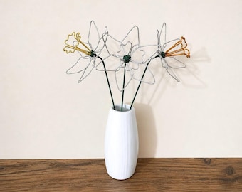May include: Three wire-sculpted flowers with silver petals and central trumpets in gold, copper, and silver tones. Each flower is attached to a thin green wire stem, and the three stems are arranged together inside a smooth, white ceramic vase.