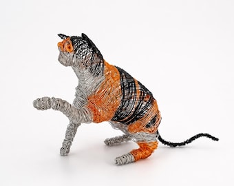 May include: A sculpture of a cat crafted from coiled wire in black, orange, and silver tones. The cat is depicted in a seated position with one front paw raised, featuring a layered wire construction that creates a textured, semi-transparent appearance.