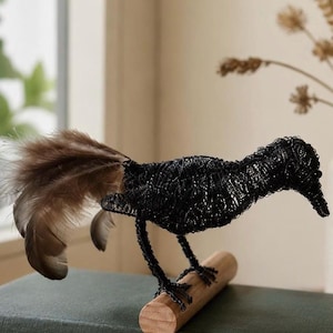May include: A black wire bird sculpture with a long beak and a tail of brown and tan feathers. The bird is perched on a small wooden dowel, with its legs and feet made of wire. The sculpture is displayed on a dark green book.