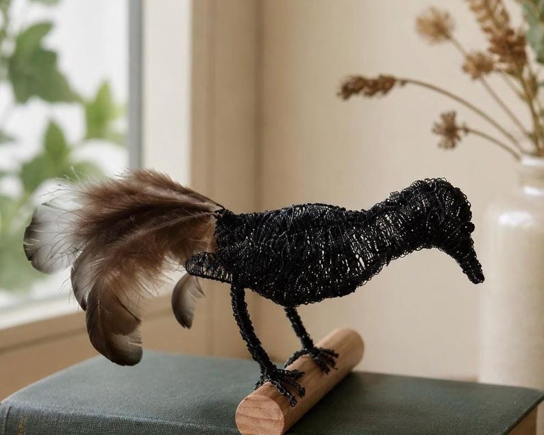 May include: A black wire bird sculpture with a long beak and a tail of brown and tan feathers. The bird is perched on a small wooden dowel, with its legs and feet made of wire. The sculpture is displayed on a dark green book.