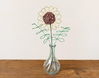 May include: A wire sculpture of a sunflower with gold petals and a dark brown center, accompanied by two green wire stems with leaves, all arranged inside a clear, ribbed glass vase with a narrow neck and a wider, rounded base.