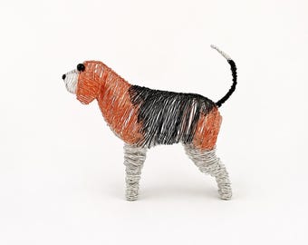 May include: A wire sculpture of a dog featuring a tri-color pattern of copper, black, and silver wire. The dog has a copper head and back, a black midsection, and silver legs and muzzle, with two small black beads serving as eyes and a tail.