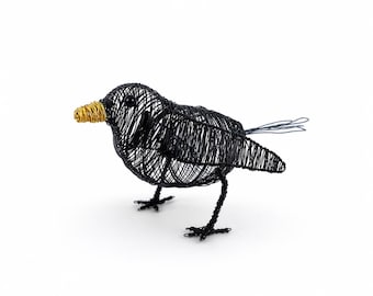 May include: A bird sculpture crafted from thin black wire, featuring a rounded body, a small black bead eye, and a distinct beak made of tightly coiled gold-colored wire. The figure stands on two thin wire legs and has a tail of loose wire strands.