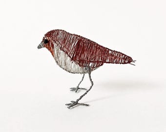 May include: A bird sculpture crafted from thin copper and silver-colored wire. The bird features a dark copper-toned back and wings, a silver-toned chest and underbelly, thin wire legs with detailed feet, and a small black bead for the eye.