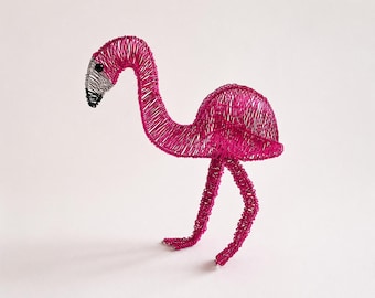 May include: A flamingo sculpture crafted from vibrant hot pink wire. The bird features a long, curved neck, a rounded body, and two thin legs. The beak is detailed with silver and black wire, and a small black bead serves as the eye.