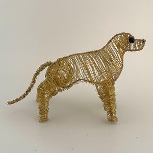 May include: A gold-coloured wire sculpture of a dog in a standing pose. The dog has a long tail and a small black bead for its eye. The sculpture is made of thin, gold-coloured wire.
