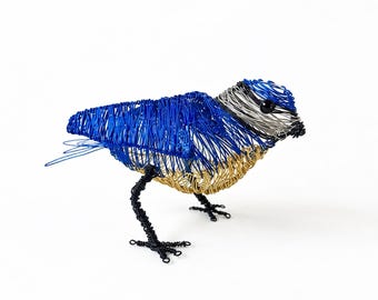 May include: A bird sculpture crafted from coiled wire, featuring a vibrant blue back and wings, a silver head with a small black bead eye, a golden yellow chest, and thin black wire legs with detailed feet, standing in a forward-leaning pose.