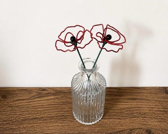May include: Two wire-sculpted red poppy flowers with black centers and green stems placed inside a clear, vertically ribbed glass bottle. The flowers feature thin, bent wire petals that form an open, organic shape, standing upright in the neck of the bottle.