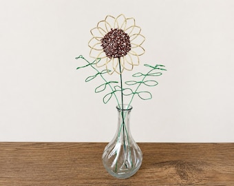May include: A wire sculpture of a sunflower with gold-colored petals and a dark brown center, accompanied by two green wire stems with leaf-shaped loops, all arranged inside a clear, ribbed glass vase with a narrow neck and a rounded base.