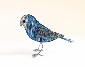 May include: A bird sculpture crafted from thin metallic wire in shades of blue and silver. The bird features a rounded body, a pointed tail, and two thin wire legs. A small, dark, circular bead serves as the eye on the side of the head.