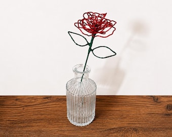 May include: A red wire rose sculpture with a green wire stem and two leaves, placed inside a clear, vertically ribbed glass bottle. The rose is constructed from coiled red wire, while the stem is wrapped in green wire.