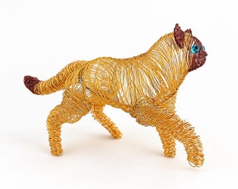 May include: A cat sculpture crafted from coiled gold-toned wire, featuring a dark brown wire face, ears, and tail tip. The feline figure has bright blue circular eyes and is depicted in a walking stance with its body angled toward the right.