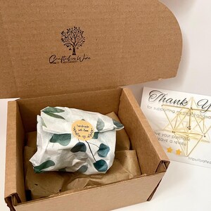 May include: A brown cardboard box with a lid, containing a wrapped gift and a thank you card. The gift is wrapped in white paper with green leaf patterns and a sticker that says "handmade with love". The thank you card has gold geometric shapes.