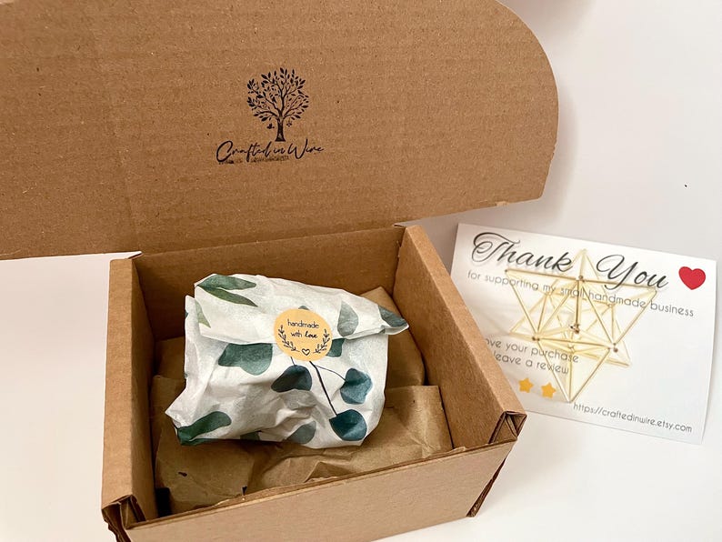 May include: A brown cardboard box with a lid, containing a wrapped gift and a thank you card. The gift is wrapped in white paper with green leaf patterns and a sticker that says "handmade with love". The thank you card has gold geometric shapes.