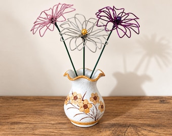 May include: A white ceramic vase with a ruffled rim and painted orange floral patterns, containing three wire-sculpted flowers in pink, silver, and purple, each featuring a central bead accent on thin green stems.