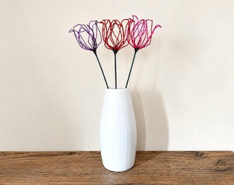 May include: A white ceramic vase with a subtle vertical ribbed texture holds three decorative wire flowers. The flowers feature open, petal-like structures in purple, red, and pink, each attached to a thin, dark green wire stem.