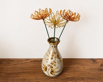 May include: A ceramic vase with a cream-colored base and brown painted leaf patterns, containing three wire-sculpted flowers. Two flowers are metallic copper-orange, and the center flower is metallic gold, all mounted on thin green stems.