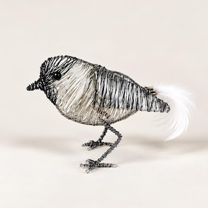 May include: A bird sculpture crafted from thin black and silver wire. The head and beak are black, while the body features a mix of silver and black wire. It has a small black eye and a fluffy white feather tail. The bird stands on two thin wire legs.