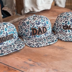 May include: Three camouflage patterned baseball caps with different names embroidered on the front. The caps are 'Riggin', 'Dad', and 'Callum'.