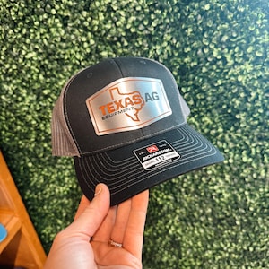 May include: Black and gray trucker hat with a silver metal patch that says "Texas Ag Equipment" with an outline of the state of Texas. The patch has a red and orange border. The hat has a black and white Richardson label on the side.