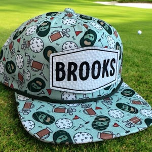 May include: A golf-themed cap with a light blue background and a pattern of golf balls, golf clubs, and beer mugs. The cap features the name "BROOKS" in bold, black letters on a white patch.