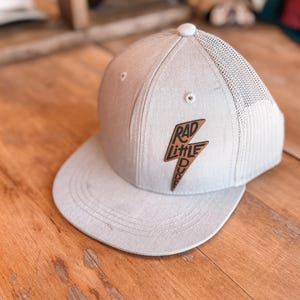 May include: A gray and black mesh trucker hat with a brown leather patch that says "Rad Little Dude" in black lettering.