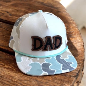 May include: A white and camouflage baseball cap with the word "DAD" in brown letters. The hat features a light blue rope detail and a camouflage pattern in shades of gray, blue, and tan. The hat is designed with a flat brim.
