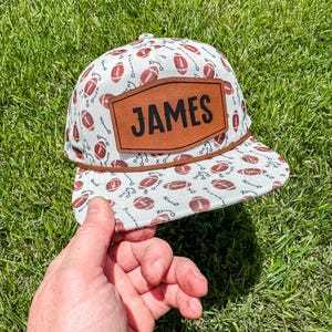 May include: A white and red football-themed cap with the name "JAMES" on a brown leather patch. The cap features a flat brim and a brown rope detail. The background is green grass.