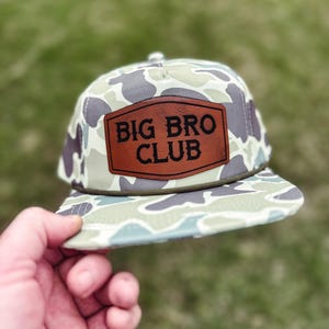 May include: A camouflage-patterned baseball cap with a brown leather patch that reads "BIG BRO CLUB." The hat has a flat brim and a snapback closure. The background is blurred green grass.